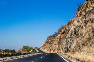 Asphalt highway with double circulation divided by a barrier with small yellow lamps, empty next to an arid rocky mountain with dry wild grass, sunny day, clear blue sky in the state of Jalisco Mexico