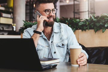 Young bearded cheerful man sits at table in front of laptops, talking on mobile phone while making notes in notebook.