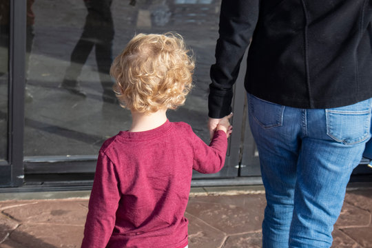 Shopping - Cropped Back View-Young Blond Curly Headed Child Holding Hand Of Mother In Jeans Walks On Sidewalk Toward Sliding Business Doors