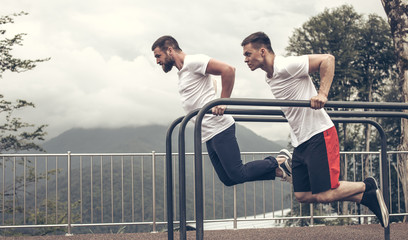 Two strong bodybuilders, athletic fitness men, with pumping up abs muscles, doing dips at outdoor...