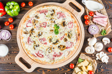 Creamy pizza with ham and mushrooms on a round cutting board on a dark wooden background. Pizza Ingredients