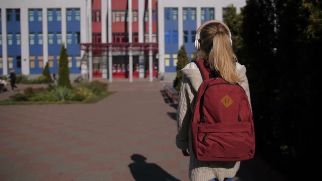 Rear view of adorable teenage girl with rucksack in headphones walking towards school building outdoors to study. Positive preadolescent schoolgirl going to classes over blurry educational building.