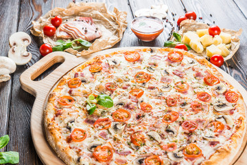 Big pizza with bacon, mushrooms and tomatoes on a round cutting board on a dark wooden background. Food Ingredients. Macro