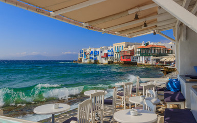 View of the famous pictorial Little Venice in Mykonos island. Splashing waves over bars and restaurants of  old town, Cyclades, Greece. Chairs with tables in typical Greek restaurant.