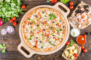 Big pizza with seafood, shrimps and olives on a round cutting board on a dark wooden background. Food Ingredients.