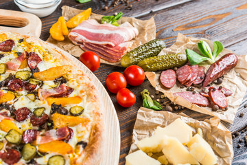 Macro. Pizza with sausage, pickled cucumber, bacon on a round cutting board on a dark wooden background. Pizza Ingredients