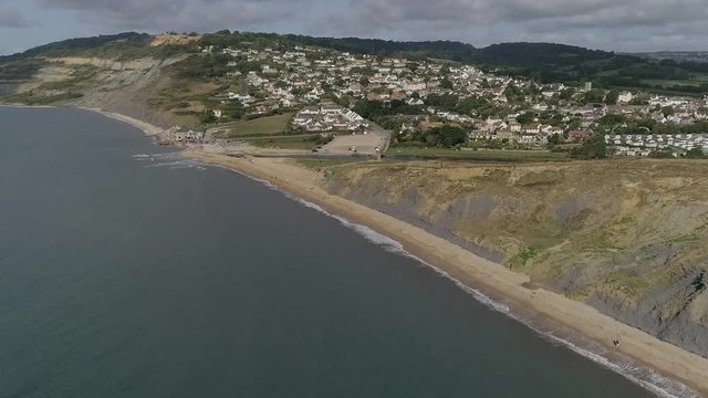 Aerial tracking from sea in towards the village of Charmouth. Amazing Jurassic coast cliffs surround the holiday destination either side. Amazing sunny day.  Vegetation in full bloom