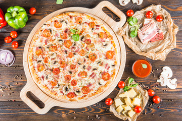 Big pizza with bacon, mushrooms and tomatoes on a round cutting board on a dark wooden background. Food Ingredients.