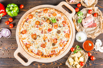 Large meat pizza with bacon, ham, chicken, sausage and tomatoes on a round cutting board on a dark wooden background. Food Ingredients.