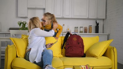 Cute schoolgirl giving her beautiful adult mother a goodbye kiss in domestic room and leaving to school. Loving mother with tablet pc sitting on sofa and saying goodbye to her girl before school.