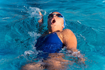 Competitive female swimmer taking big breath of air as she speeds toward finish line during backstroke race. - obrazy, fototapety, plakaty