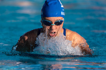 Pool water illuminated by afternoon sun cascades of the face of female swimmer as she comes up for air during breast stroke. - obrazy, fototapety, plakaty