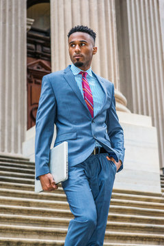 Young African American Businessman with beard working in New York, wearing dark sky blue suit, white striped shirt, violet red patterned tie, walking down stairs from old style office building.