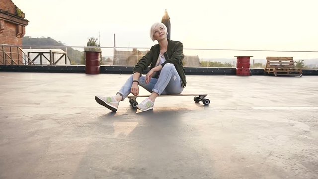 Cute teen girl is sitting on her skateboard of the rooftop modern building. Young blonde woman sitting on longboarding at sunrise at the city