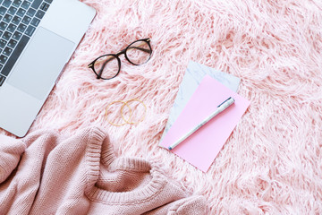 Flatlay of laptop, women knitted sweater, reading glasses, paper, pen on a pink fur background