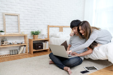 Young asian couple looking at laptop computer to search for travel plan, book hotel room, buy ticket, purchase travel insurance or shopping online together at bedroom in e-commerce technology concept.