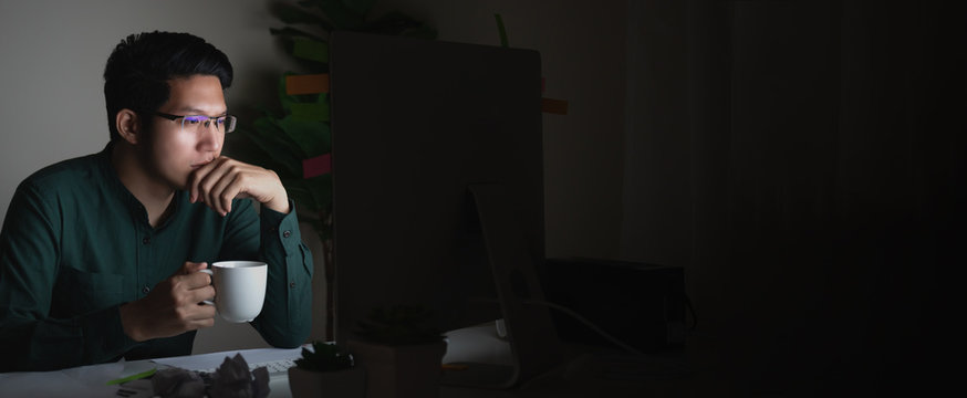 Banner Of Attractive Young Asian Man Drinking Coffee Sitting On Desk Table Looking At Laptop Computer In Dark Late Night Working Feeling Serious Thinking And Determinated At Home Office Concept.