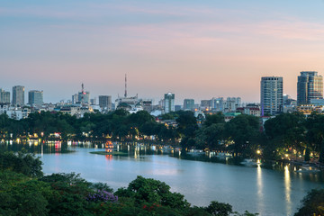 Fototapeta na wymiar Hoan Kiem lake or Sword lake, Ho Guom in Hanoi, Vietnam with Turtle Tower, green trees and buildings on horizon, at twilight period