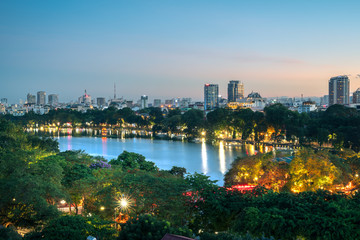Fototapeta na wymiar Hoan Kiem lake or Sword lake, Ho Guom in Hanoi, Vietnam with Turtle Tower, green trees and buildings on horizon, at twilight period