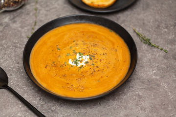 Cream soup of carrots and pumpkin with herbs, in a black bowl on a gray background.