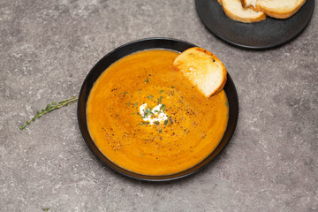 Cream soup of carrots and pumpkin with herbs, in a black bowl on a gray background.