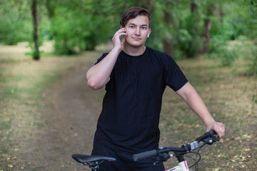 Attractive young caucasian man touches his white earphones holding the bicycle, standing at the abandoned park. Dark casual wear, dark hair and eyes. Outdoors, close up, copy space.