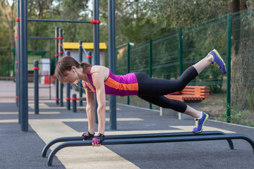 Young caucasian woman workouts on the park sports ground. In sports plank position with one leg up, sportswear. White earphones, protective gloves. Real woman in morning excersice. Copy space, outdoor