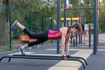 Young caucasian woman workouts on the park sports ground. In sports plank position with one leg up,...