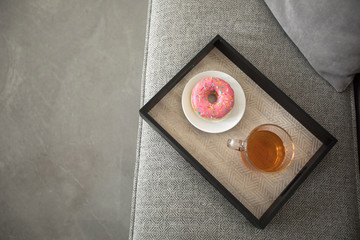 From above shot of tray with served cup of fresh tea and pink glazed doughnut on gray sofa