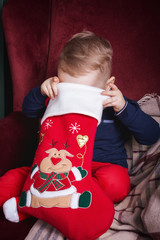 Cute lovely small boy holding a red christmas stocking sitting on the classic velvet red armchair