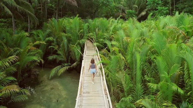 Aerial view of woman walking on long wooden bridge holding selfie stick among palm trees in Bojo river, Philippines.