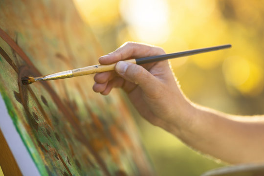 artist drawing a picture on an easel in nature, a hand makes a brush stroke on a canvas, a concept of creativity and a hobby