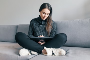 Autumn winter portrait of a young girl in a warm knitted sweater at home on the sofa with a digital...