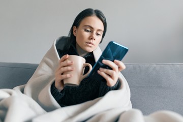 Autumn winter portrait of a young girl resting at home on the sofa with mobile phone and cup of hot drink, under warm blanket