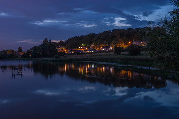 A Well Lit Village Viewed Across Water Against An Evening Sky, Fairburn, West Yorkshire