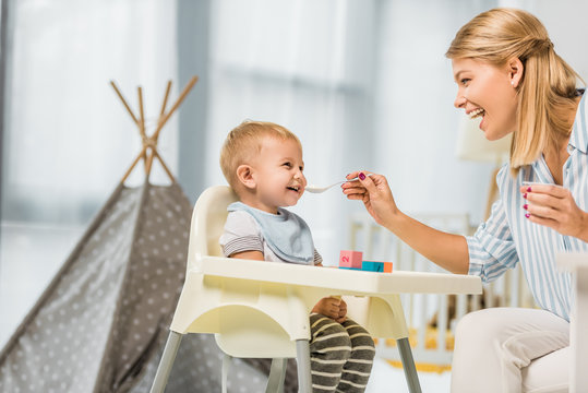 cheerful mom feeding son in highchair with baby food in nursery room