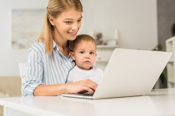 mother sitting at desk with cute toddler while using laptop