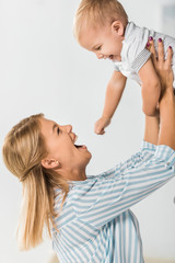 cheerful mother looking at toddler and holding him in hands on white background