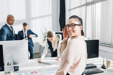 attractive young businesswoman in eyeglasses smiling at camera while working with colleagues in office