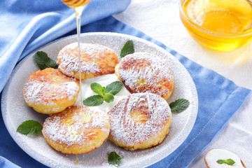 Cottage cheese pancakes sprinkled powdered sugar and fresh mint. Cheesecakes on white plate. Sour cream in wooden spoon. Honey and dairy products on white blue background. Healthy food
