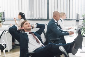 smiling young businessman sitting with hands behind head and looking up in office