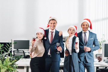 happy young multiethnic coworkers holding glasses of wine and smiling at camera while celebrating new year in office