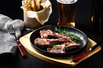 Grilled lamb chops in a cast-iron frying pan with fried potatoes and glass of beer on dark background