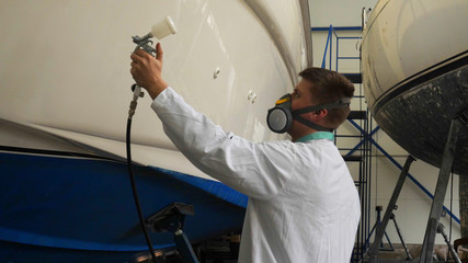 A young man (guy) is a specialist in cleaning the yacht, in a respirator and using a spray on the background of the yachts. Concept from: Professional, Specialist, Cleaning, Yacht service, Preparation