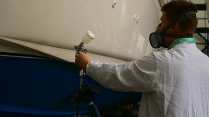A young man (guy) is a specialist in cleaning the yacht, in a respirator and using a spray on the background of the yachts. Concept from: Professional, Specialist, Cleaning, Yacht service, Preparation