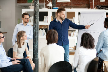 Millennial male mentor or coach make flipchart presentation to diverse colleagues at meeting,...