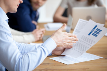 Close up of male employee read paper handout material during office meeting, man analyzing paperwork report at briefing, explaining content to colleague, worker sit at desk looking through document