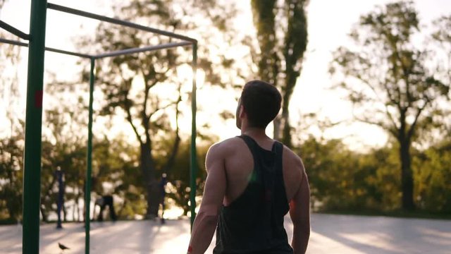 Fit man in black shirt doing pull-ups on horizon bar on sports ground with trees and sun shines on the background