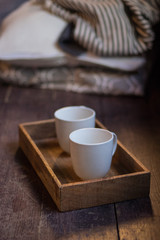 Two white porcelain teacups in wooden tray with cozy pillow and blanket . Selected focus