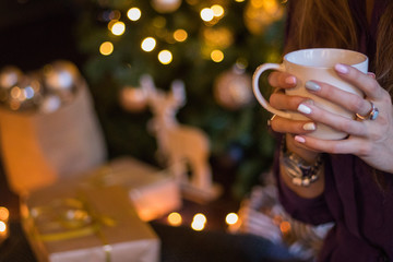 Young girl sitting near the Christmas tree with gifts and drinking coffee from a white cup. Selected focus